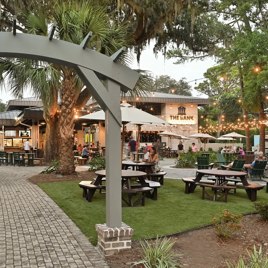Outdoor seating area with picnic tables, string lights, and people near The Bank restaurant at dusk.