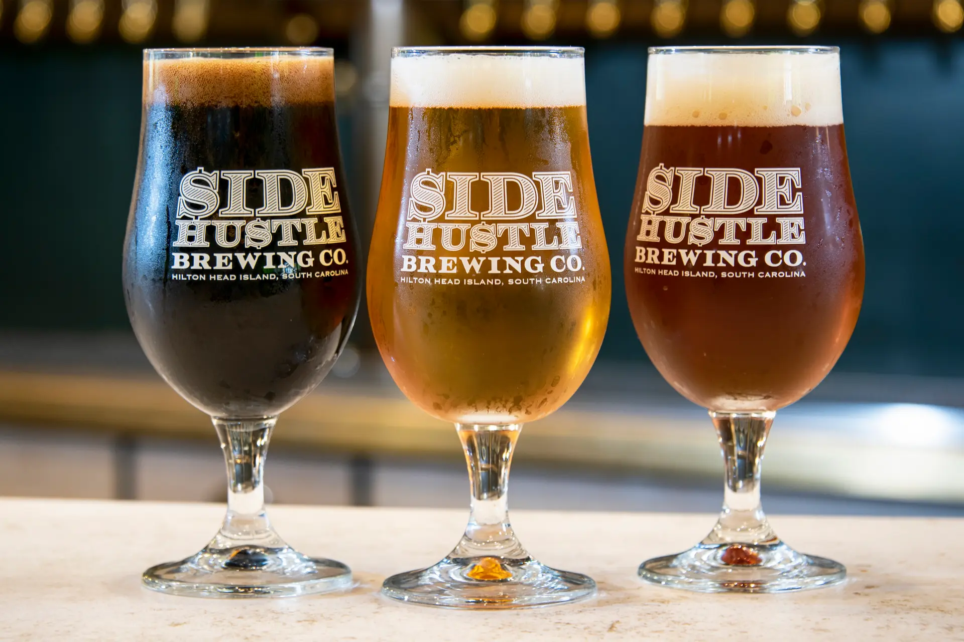Three side hustle brewing co beers in glasses: dark stout, golden lager, and amber ale.