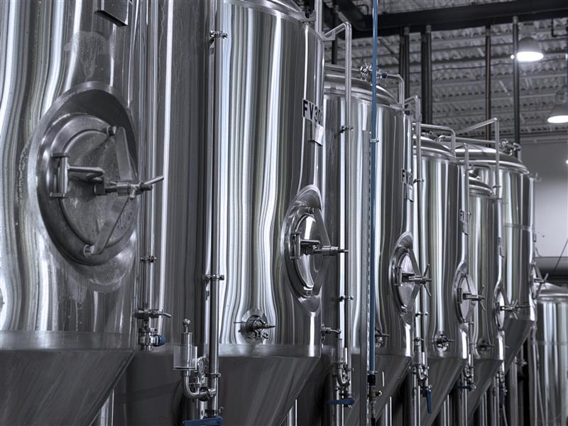 Row of large stainless steel brewing tanks with pipes in a brewery facility.