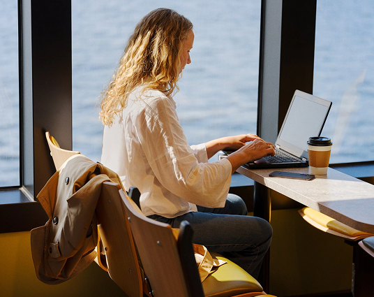 Woman with curly blonde hair working on a laptop at a window-side table with a coffee cup and a phone nearby.