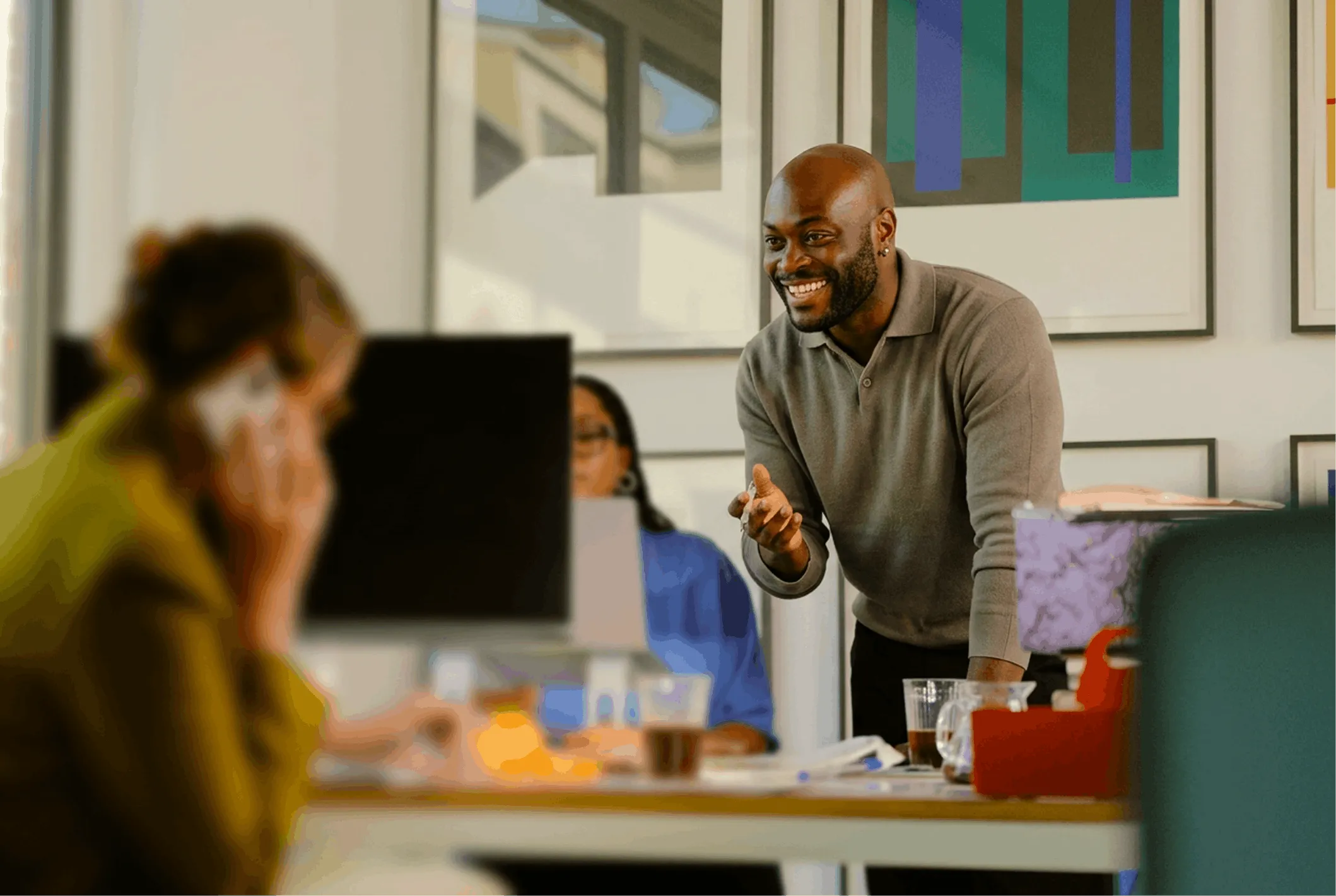 Smiling man in a gray sweater leaning over a desk and engaging with colleagues in a bright office.