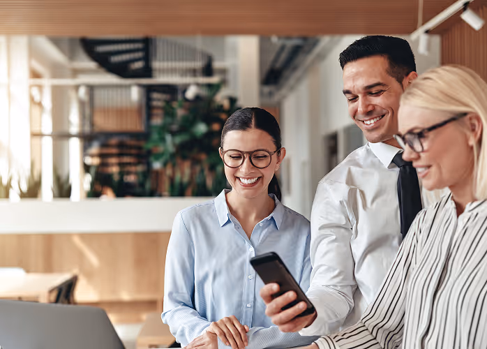 Three business professionals smiling and looking at a smartphone in a modern office setting.
