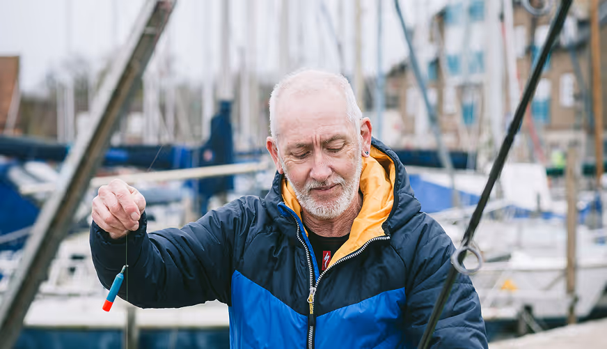 Older man in a blue and black jacket holding a fishing bobber by a marina with boats in the background.