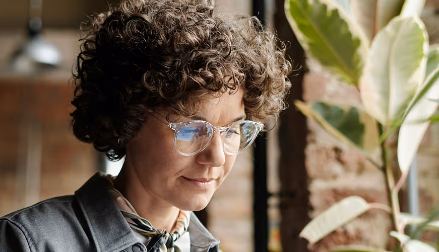 Person with curly hair and clear glasses looking down thoughtfully indoors near a plant and brick wall.