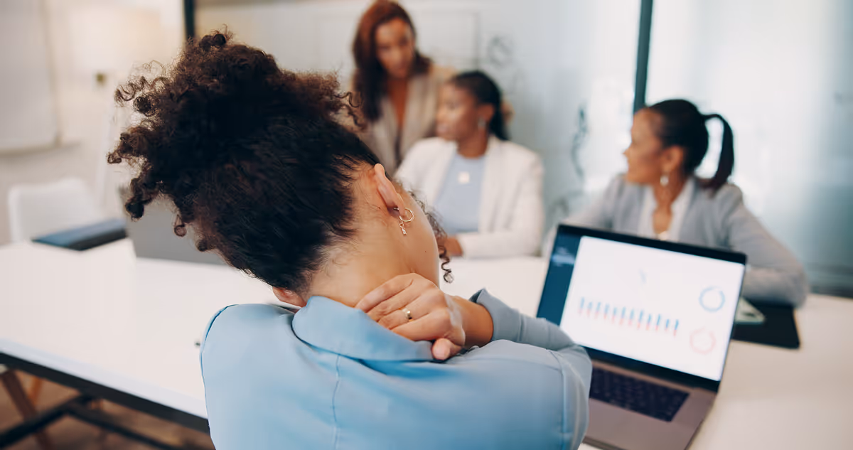 Woman in blue shirt holding her neck in pain sitting by a laptop with colleagues in a meeting room.