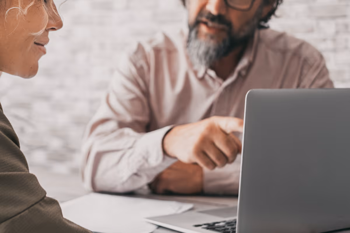 Man with beard and glasses pointing at laptop screen while woman listens attentively during discussion.