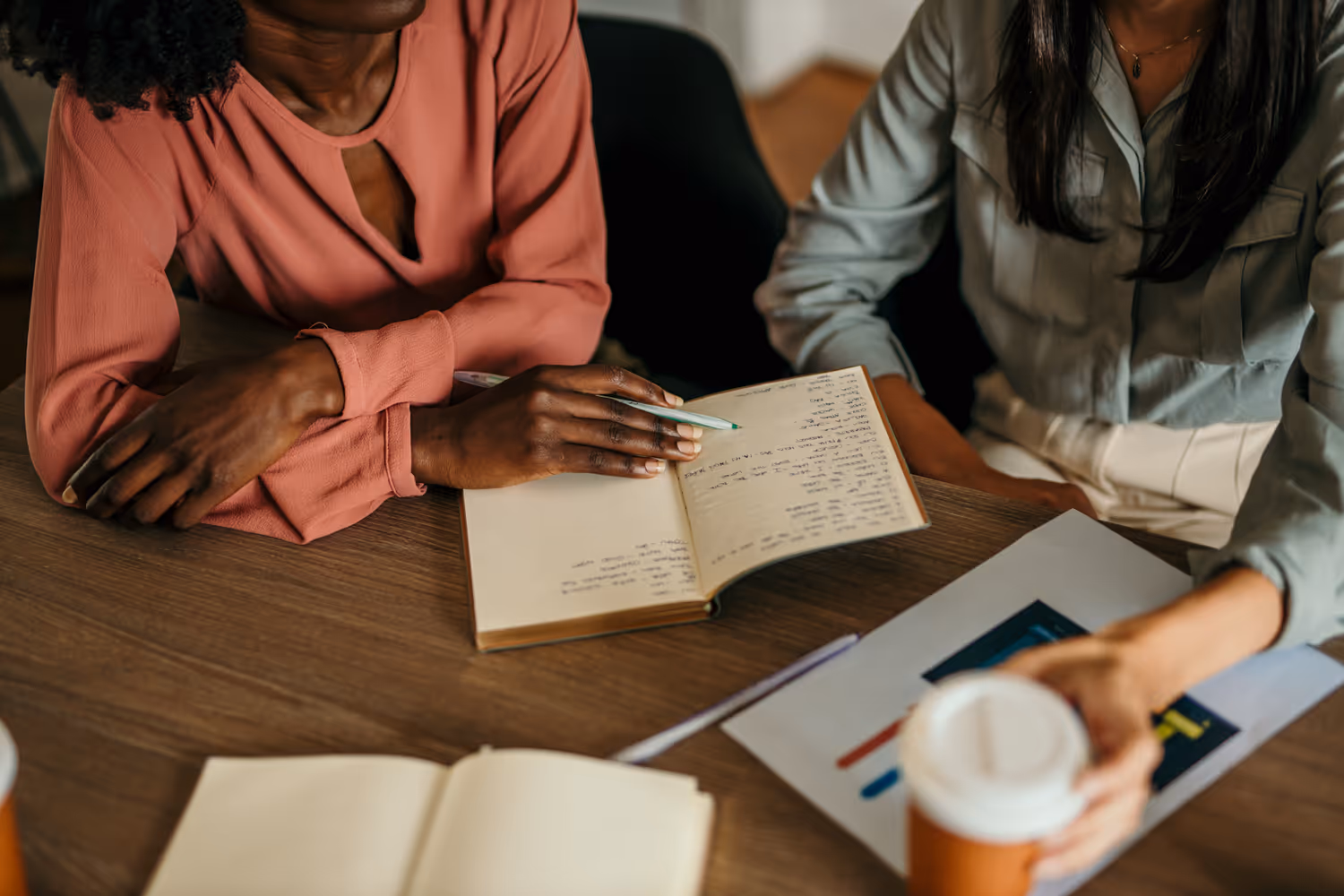 Two women sitting at a table, one writing in a notebook, the other reaching for a takeaway coffee cup.