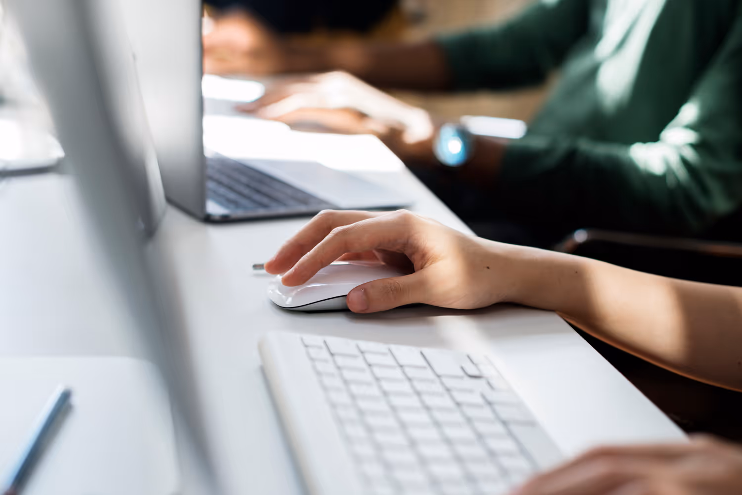 Person's hand using a white computer mouse next to a keyboard and laptop on a desk.