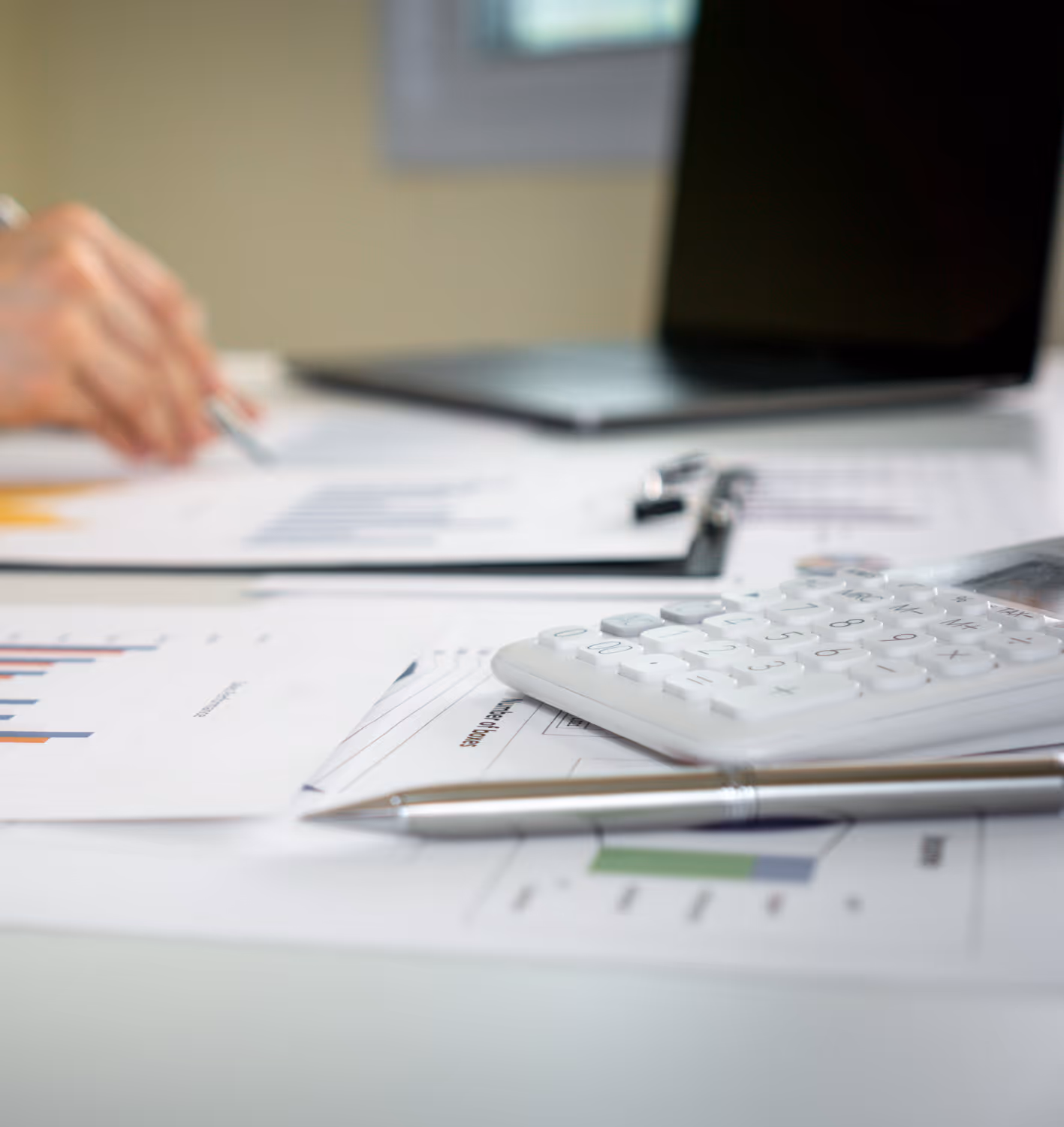 Calculator, pen, financial papers, and laptop on desk with hand holding pen in background.