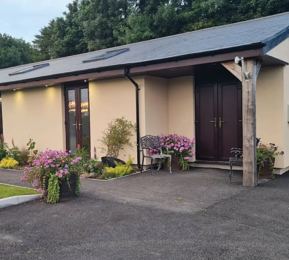 Modern beige building with two wooden double doors, skylights on the roof, metal chairs, and flower pots with pink and purple blooms.