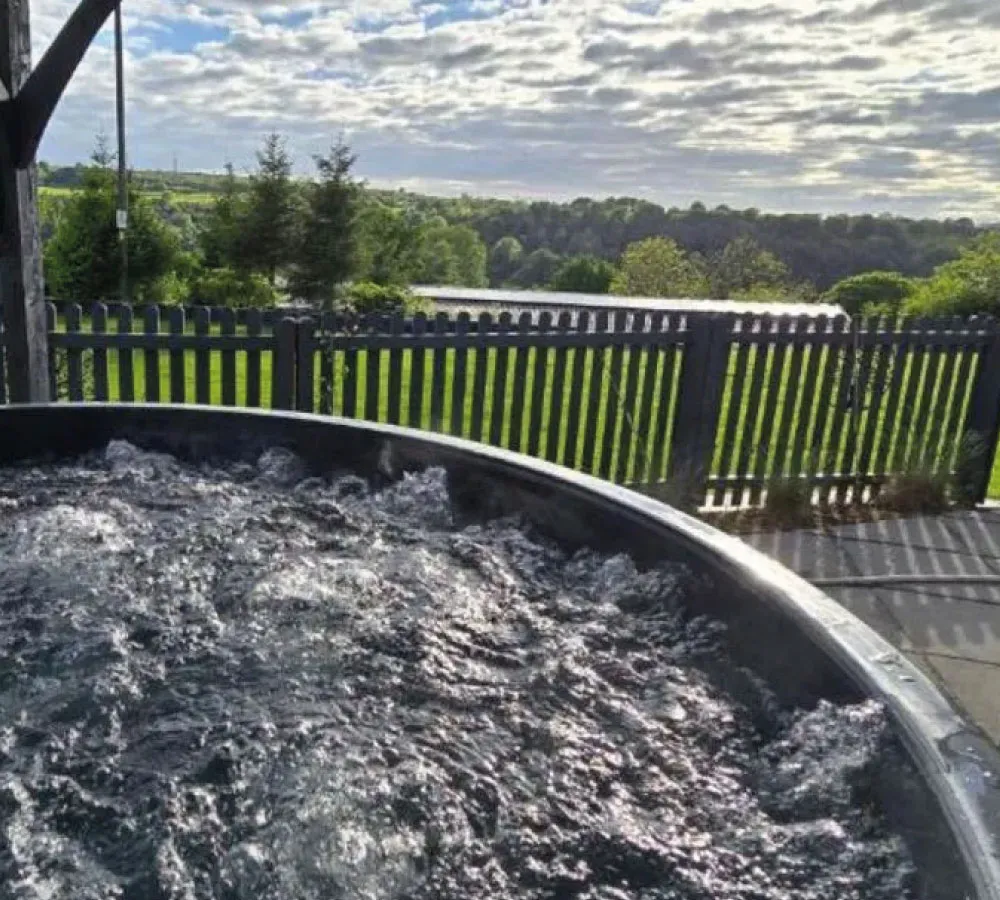 Bubbling water in a hot tub with a wooden fence and green trees under a partly cloudy sky in the background.