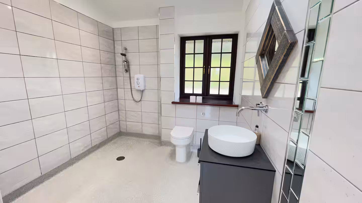 Modern bathroom with a walk-in shower, white toilet, round white sink on dark vanity, and window with wooden frame.