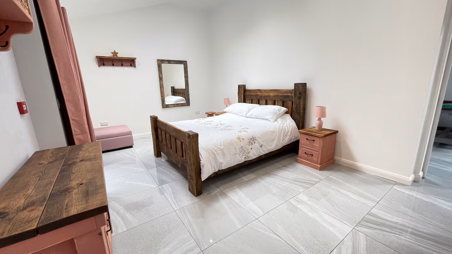 Bedroom with a wooden bed frame, white bedding with floral patterns, pink bedside table with a lamp, a wall mirror, pink ottoman, and tiled floor.