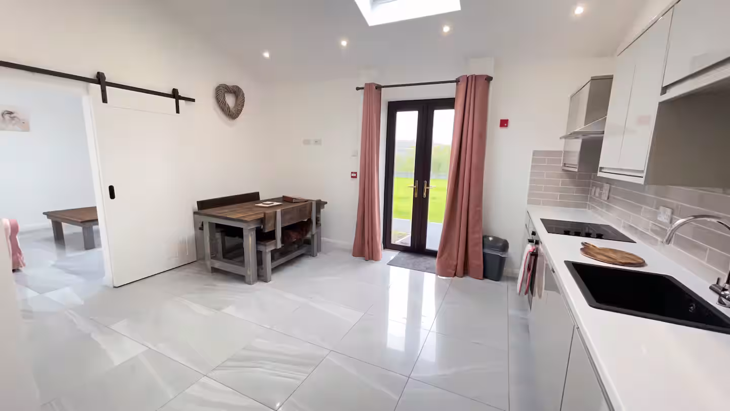 Modern kitchen with white cabinets, gray tiled backsplash, black sink, and stove, a wooden dining table with benches, pink curtains on glass doors, and a view into an adjoining room with a wooden table.