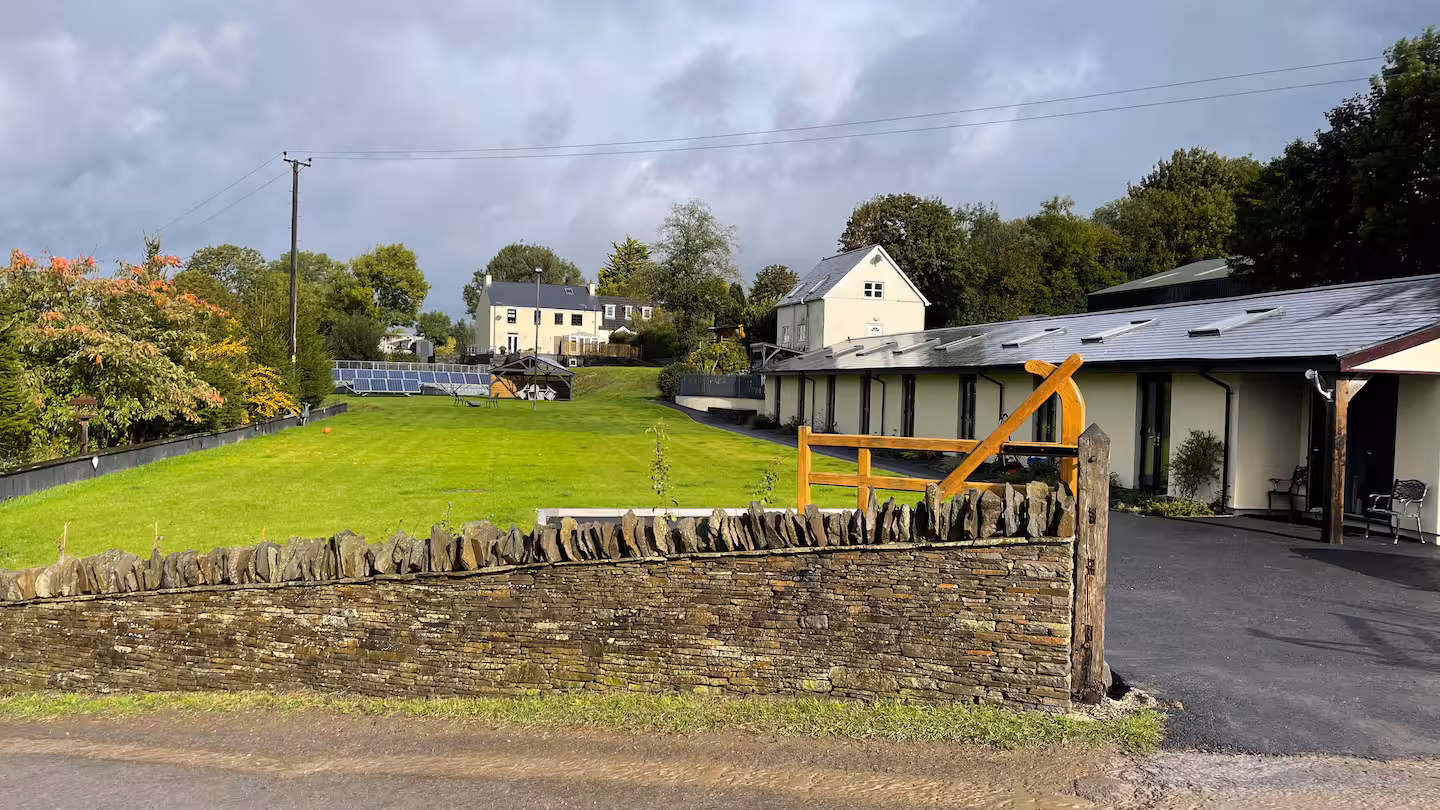 A rural scene with stone wall and wooden gate in foreground, green lawn with solar panels, and houses surrounded by trees under a cloudy sky.