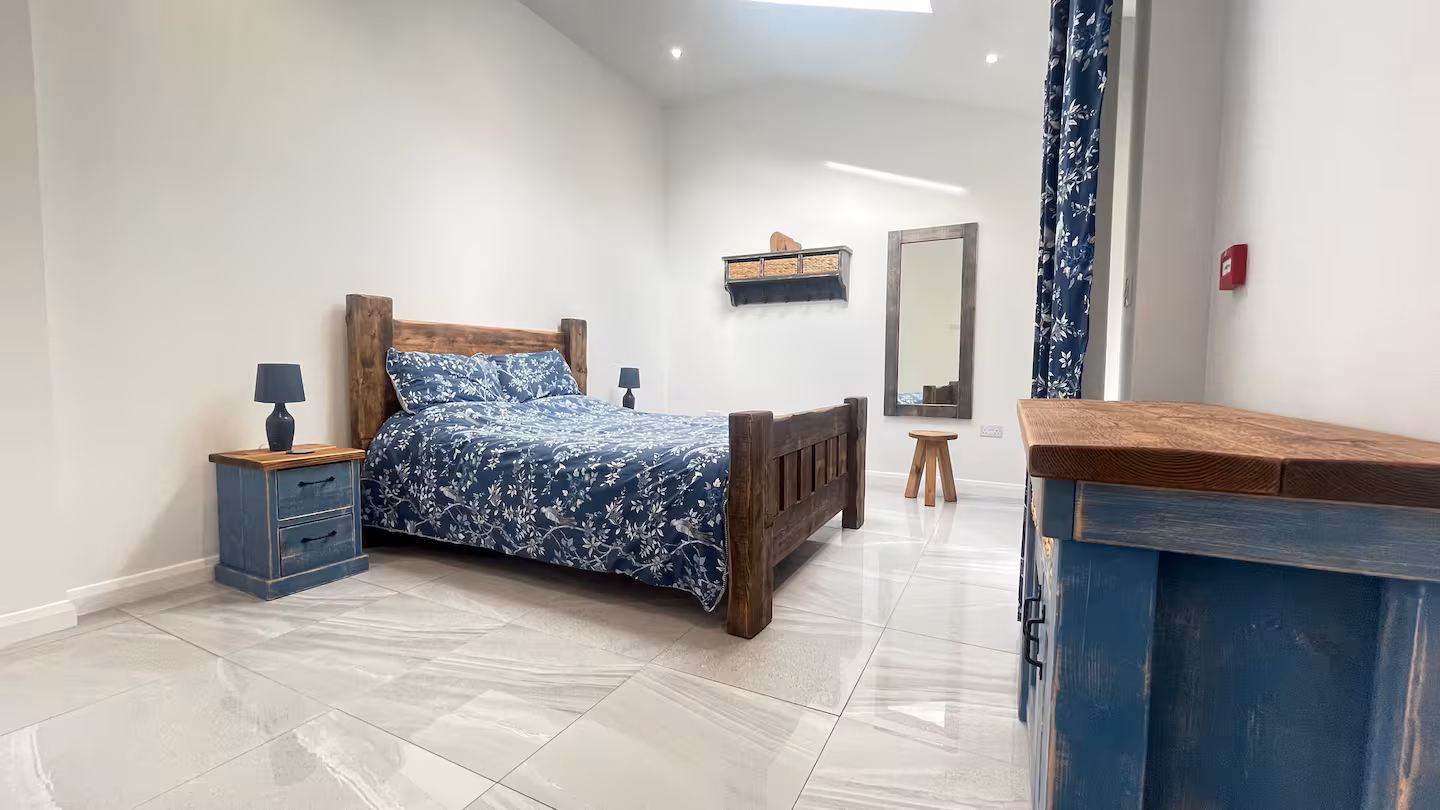 Minimalist bedroom with wooden bed frame, blue floral bedding, matching nightstand and dresser, a wall mirror, and white tiled floor.