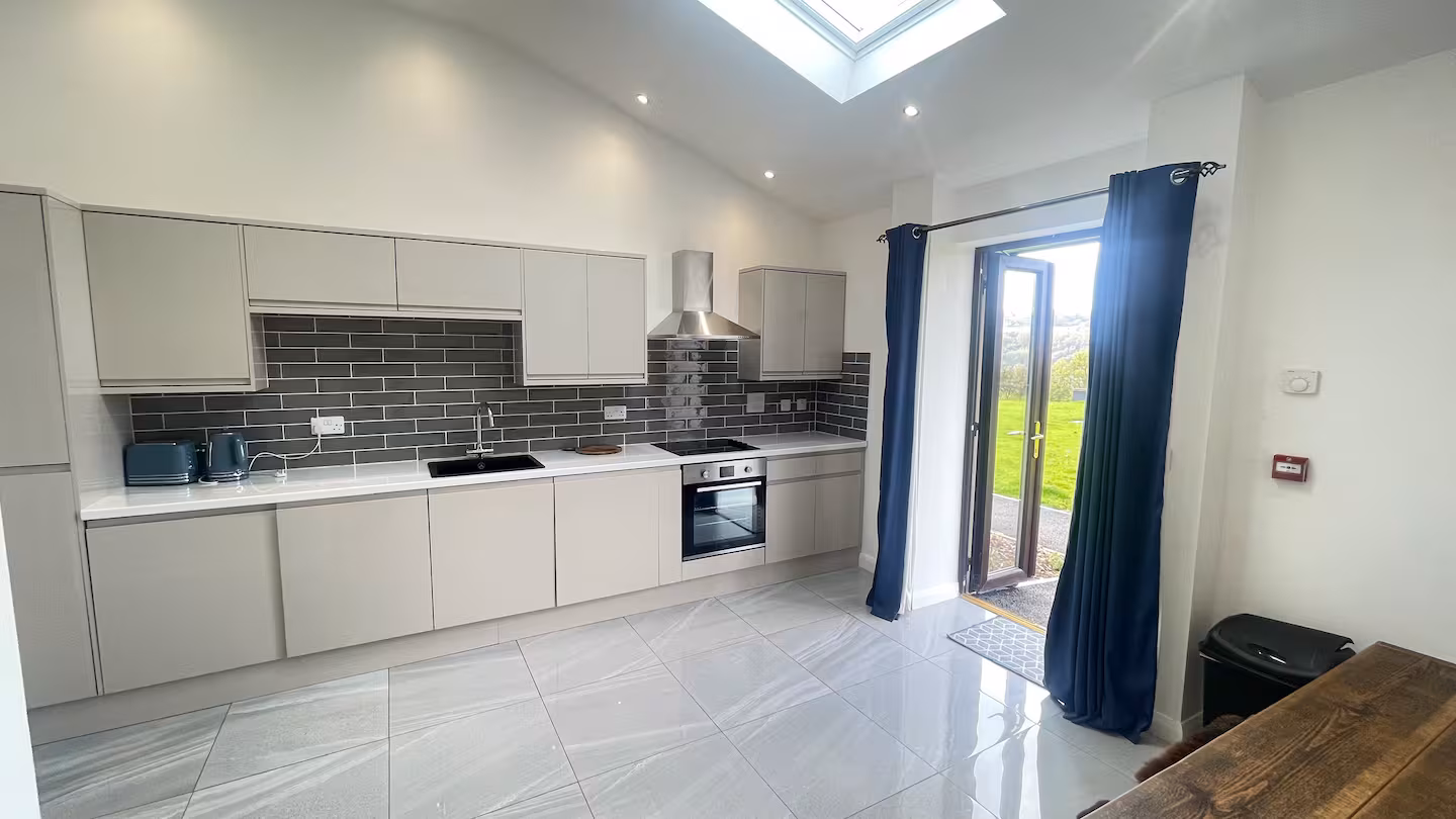 Modern kitchen with light gray cabinets, black tile backsplash, stainless steel oven and hood, blue toaster and kettle, and glass door opening to a green outdoor area.