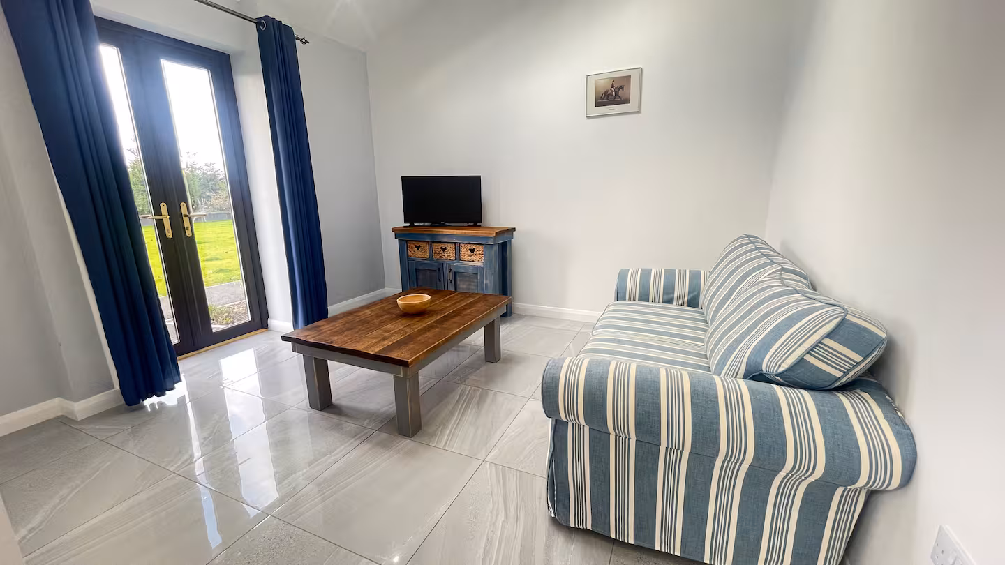 Living room with a blue and white striped sofa, wooden coffee table with a bowl, blue cabinet with a TV, and glass doors with blue curtains opening to a garden.