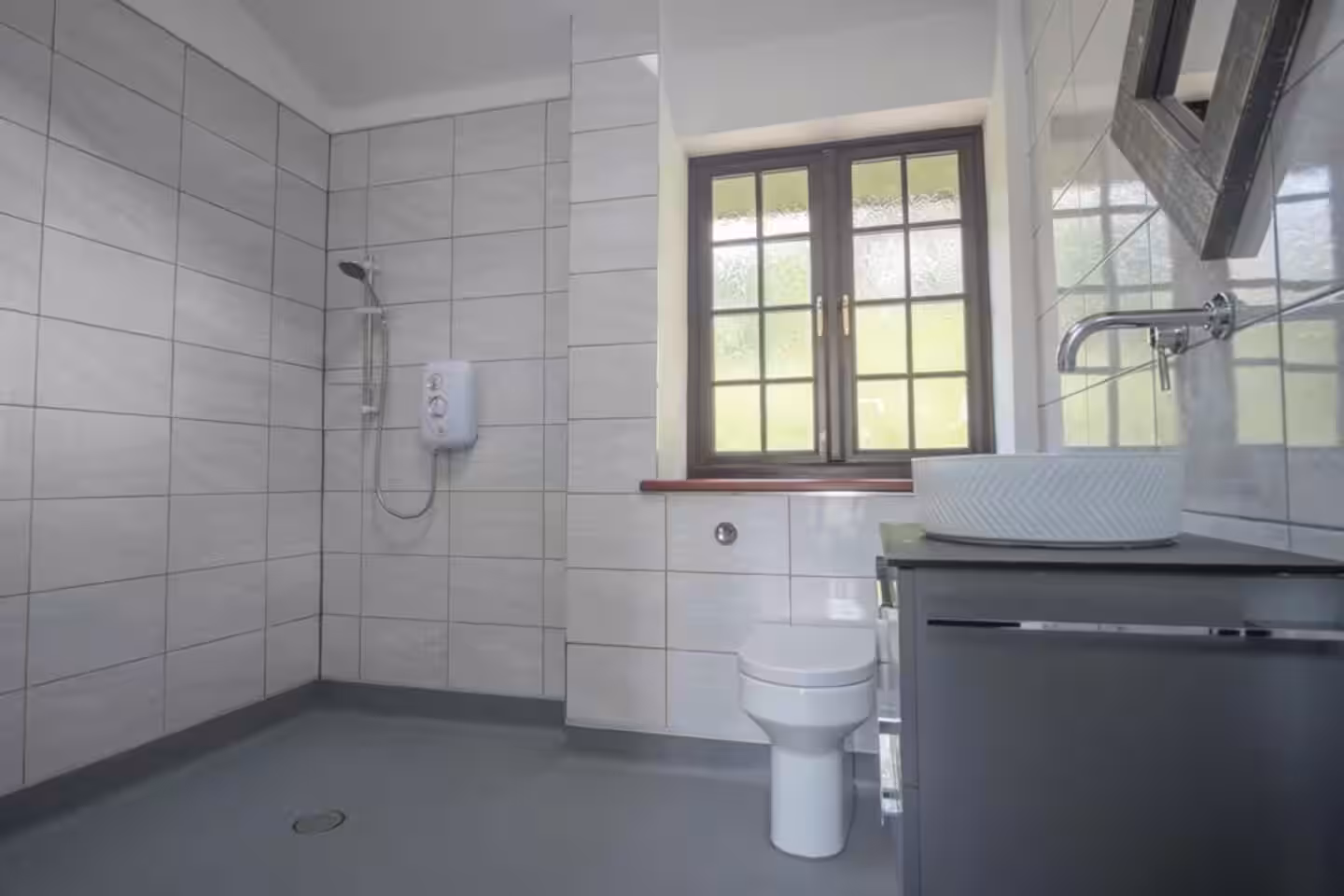 Modern bathroom with a tiled walk-in shower, white toilet, and a black vanity with a round sink under a window.