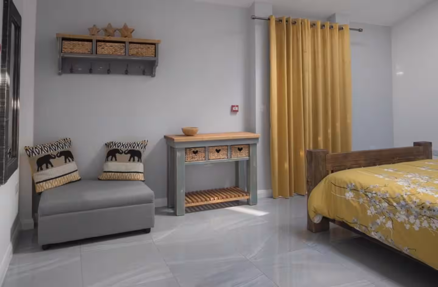 Cozy bedroom corner with a wooden bed with yellow floral bedding, mustard yellow curtains, a gray ottoman with elephant-patterned cushions, and a small wooden console table with woven drawers.