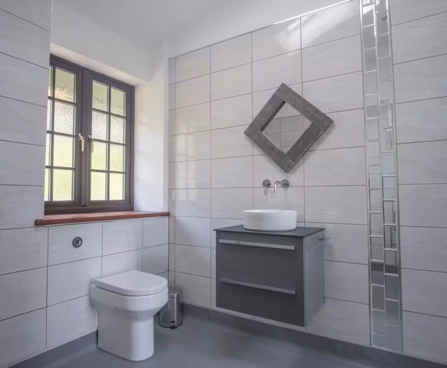 Modern bathroom with white tiled walls, a square toilet under a window, and a wall-mounted gray vanity with a white round vessel sink and a diamond-shaped mirror above.