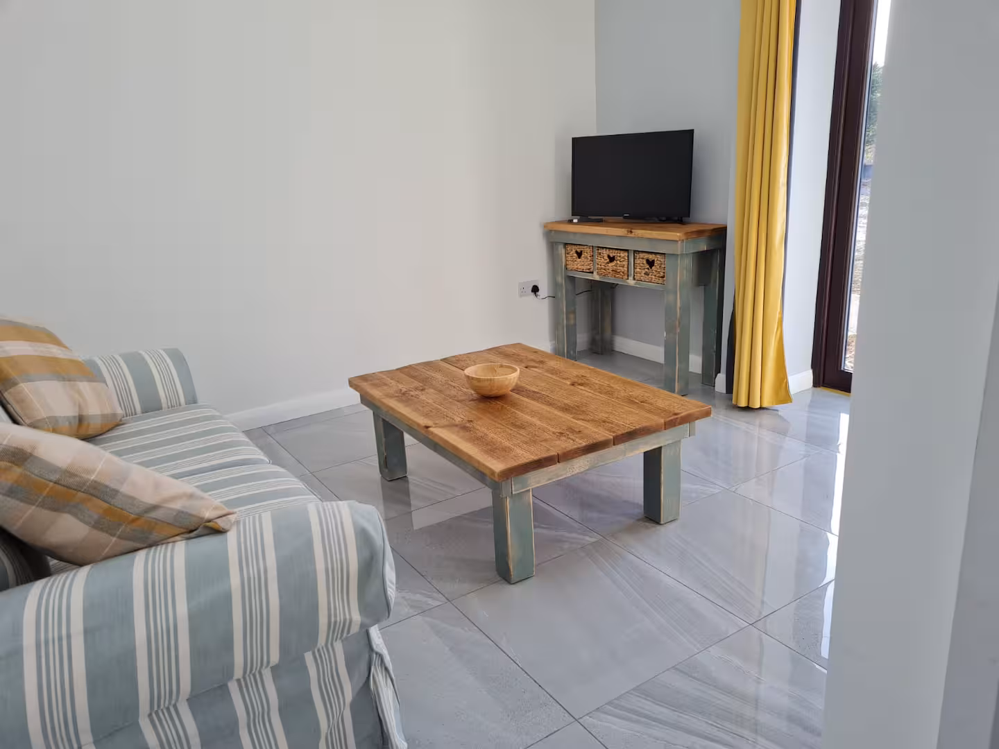 Small living room with striped sofa, wooden coffee table with bowl, TV on console table, and yellow curtains by glass door.