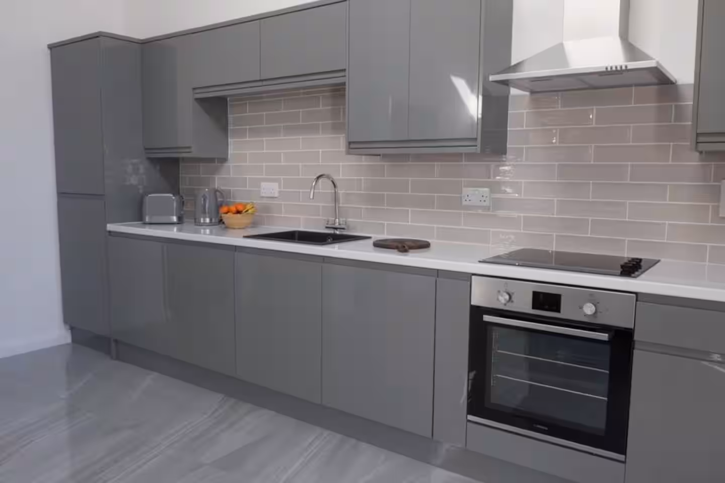 Modern kitchen with gray cabinets, white countertop, built-in oven, stovetop, stainless steel range hood, and a sink with a chrome faucet.