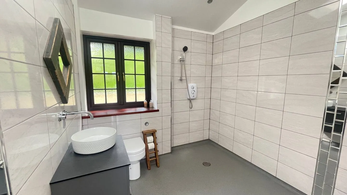 Modern bathroom with white tiled walls, a wall-mounted faucet above a round white sink on a dark vanity, a white toilet, a wooden toilet paper holder, and a walk-in shower with a wall-mounted showerhead.