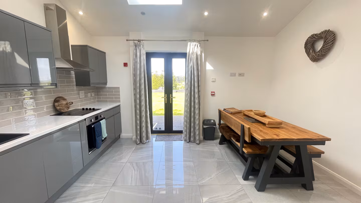 Modern kitchen with gray cabinets, stainless steel oven and hood, large tiled floor, and wooden dining table with benches under natural light.