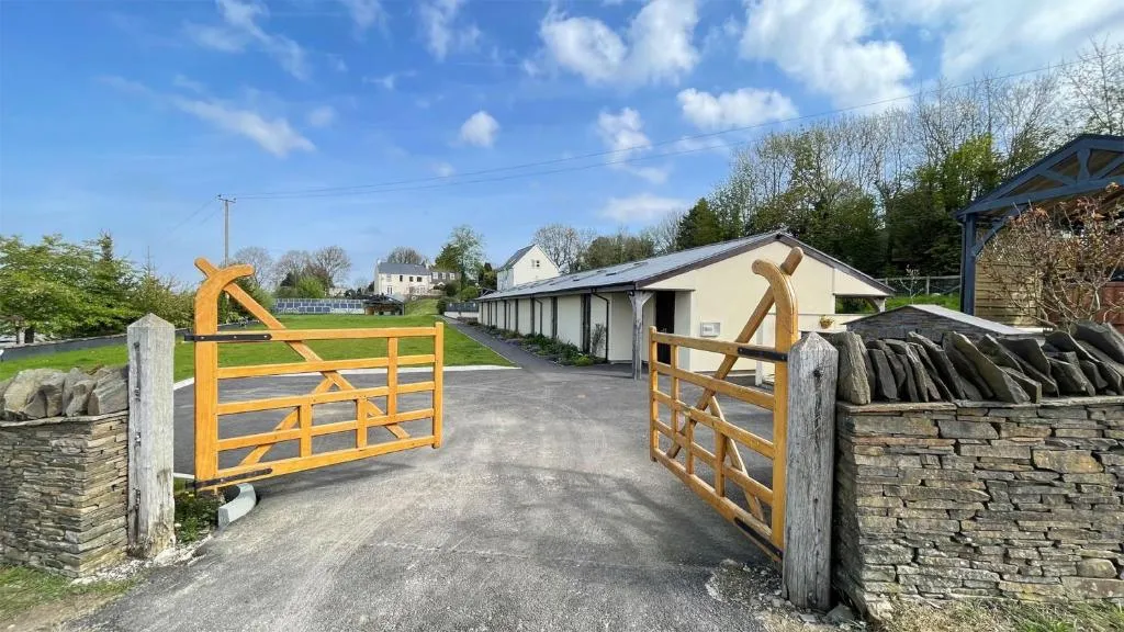 Open wooden farm gate leading to a paved driveway with buildings and houses under a partly cloudy blue sky.