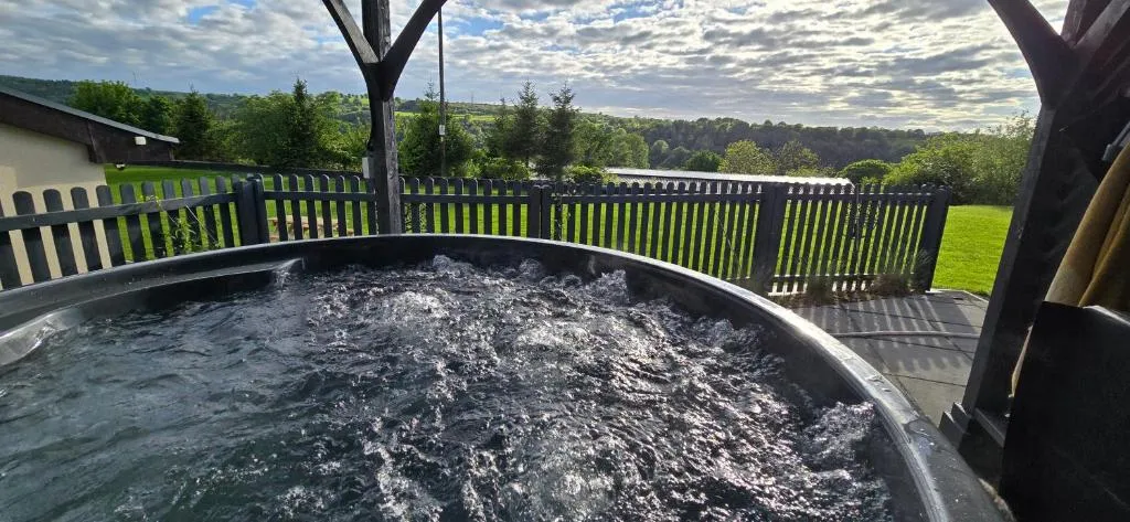 Bubbling hot tub under a wooden pergola overlooking a fenced grassy yard and distant tree-covered hills under a cloudy sky.