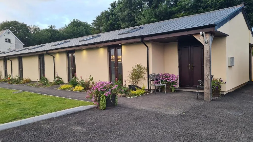 Single-story beige building with dark brown doors and windows, surrounded by a pathway, flower pots with pink flowers, and greenery.