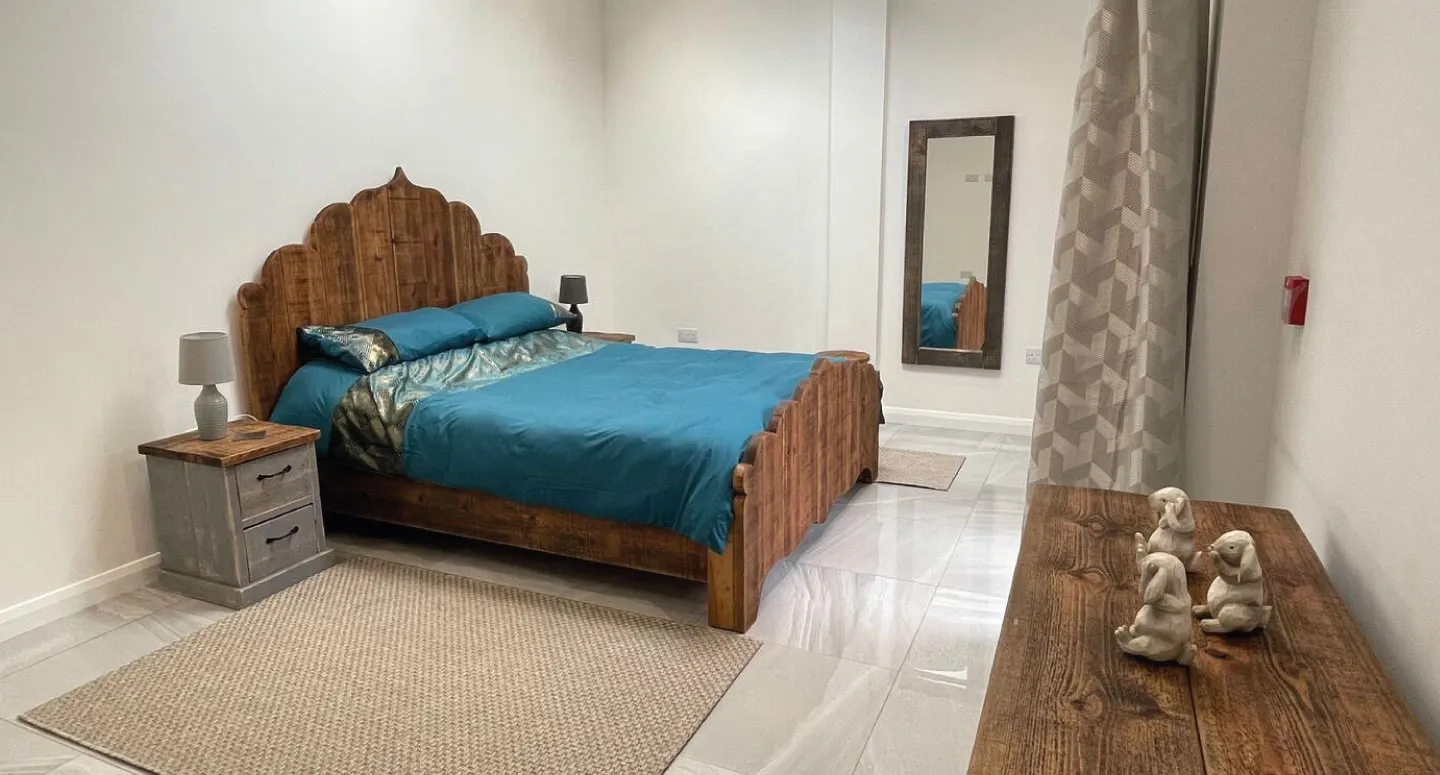 Bedroom with wooden bed frame and blue bedding, a nightstand with lamp, a full-length mirror, patterned curtain, and three small white animal figurines on a wooden dresser.