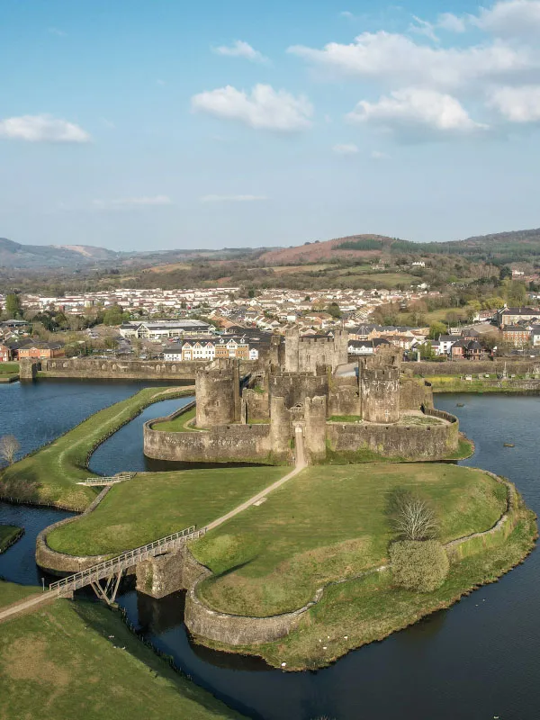 Aerial view of a medieval stone castle surrounded by a moat with a bridge, green grassy area, and town in the background under a partly cloudy sky.