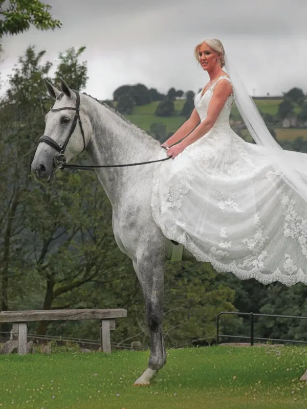 Bride in a white lace wedding dress sitting on a gray horse outdoors with trees and hills in the background.