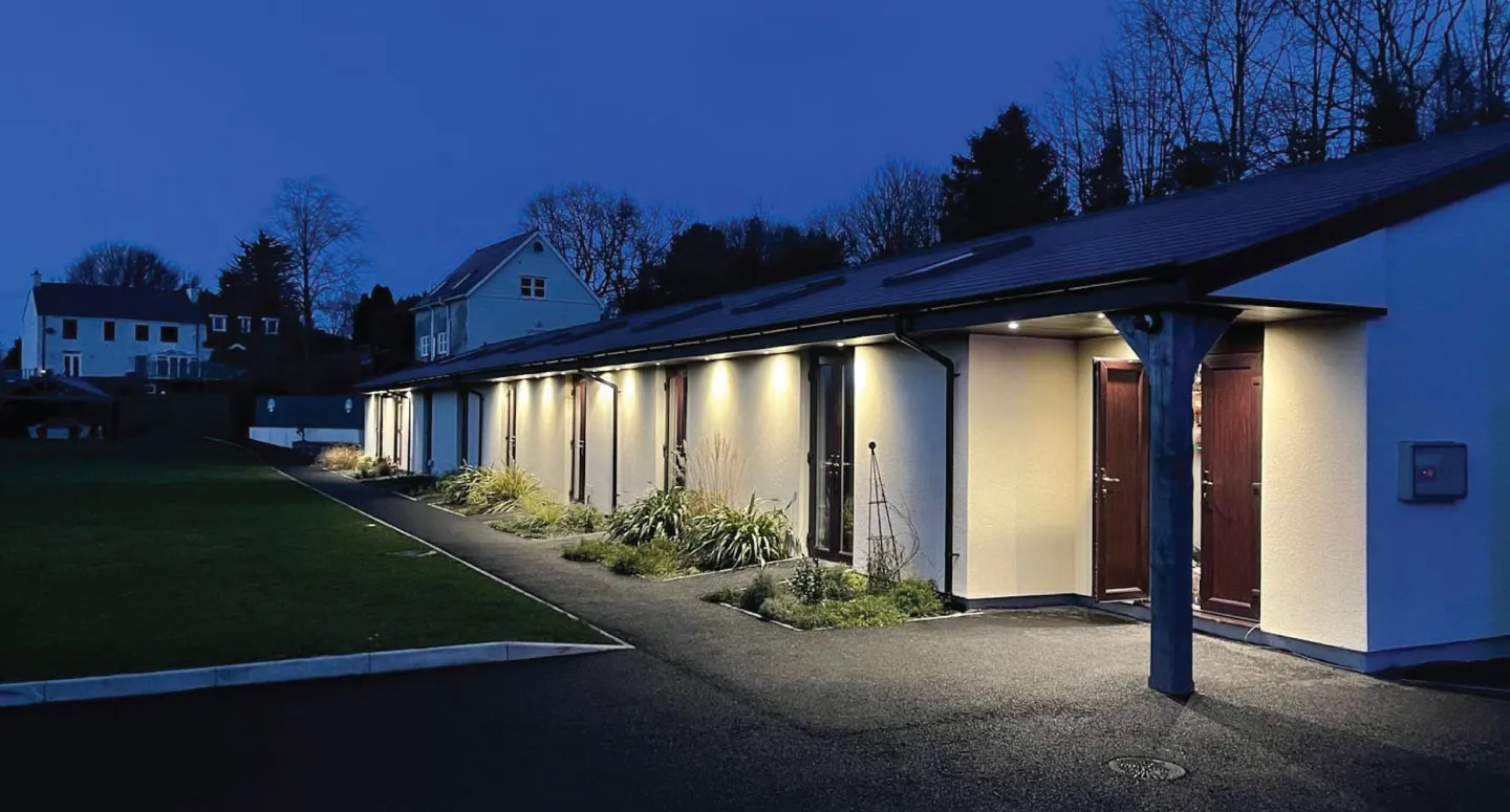 Row of modern single-story rooms with exterior lighting at dusk and a pathway bordered by grass and plants.