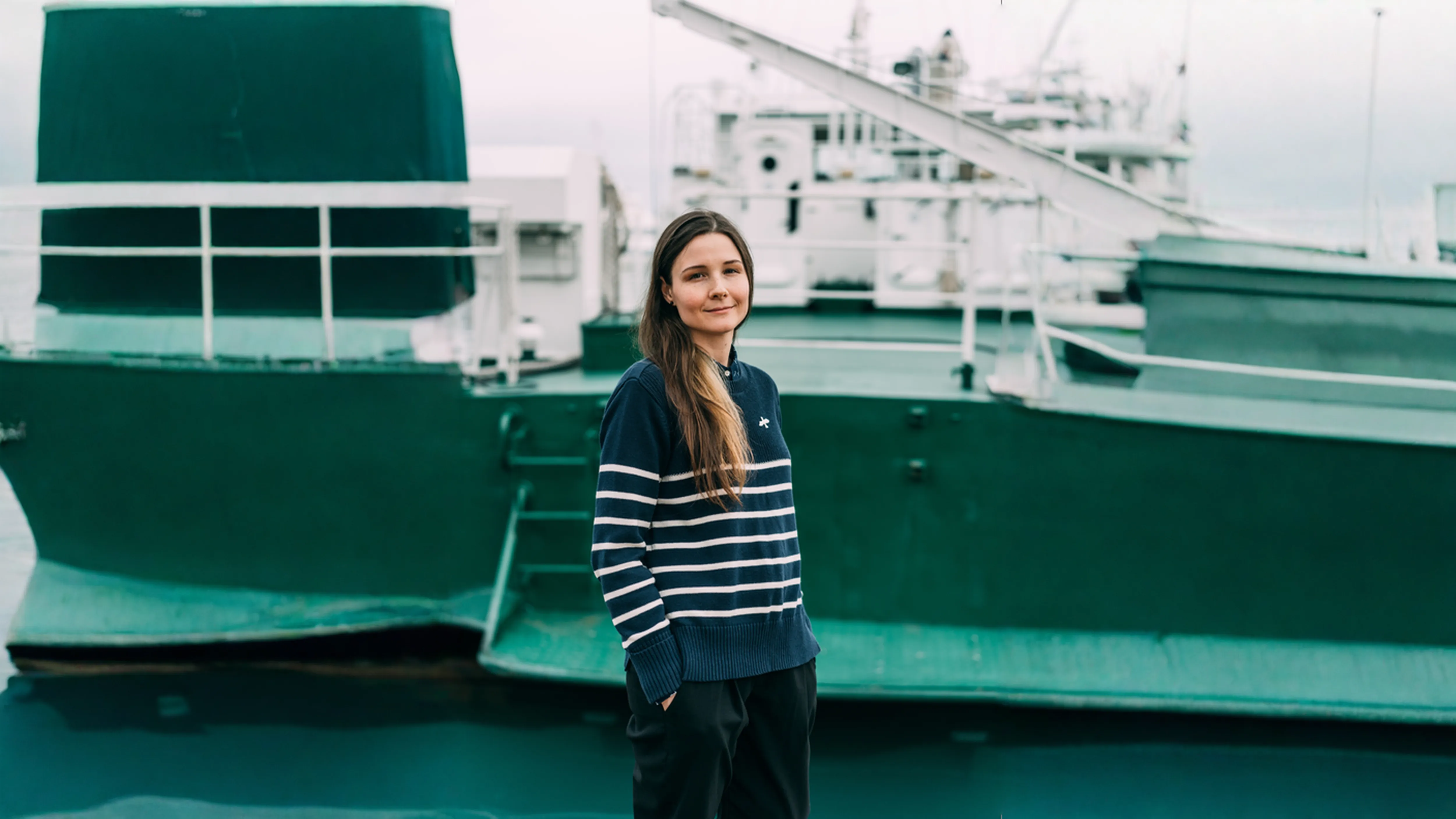 Frau mit gestreiftem Pullover und langen Haaren steht vor einem grünen Schiff am Wasser.