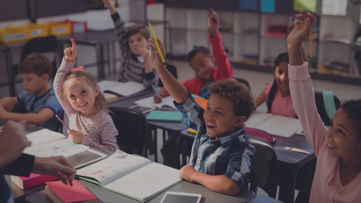 Élèves d'une classe d'école primaire levant la main pour répondre, cahiers et tablettes sur les tables