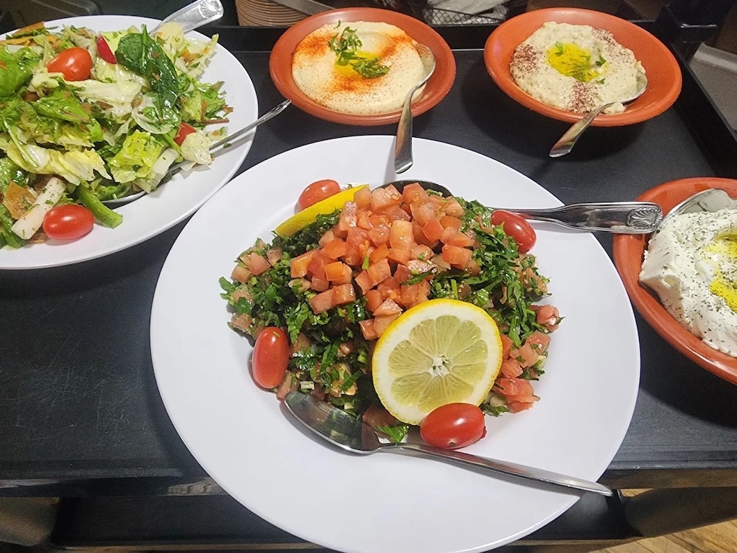 Plates of fresh Mediterranean dishes including tabbouleh salad with tomatoes and lemon, leafy salad, hummus, and a white creamy dip with olive oil on a black table.