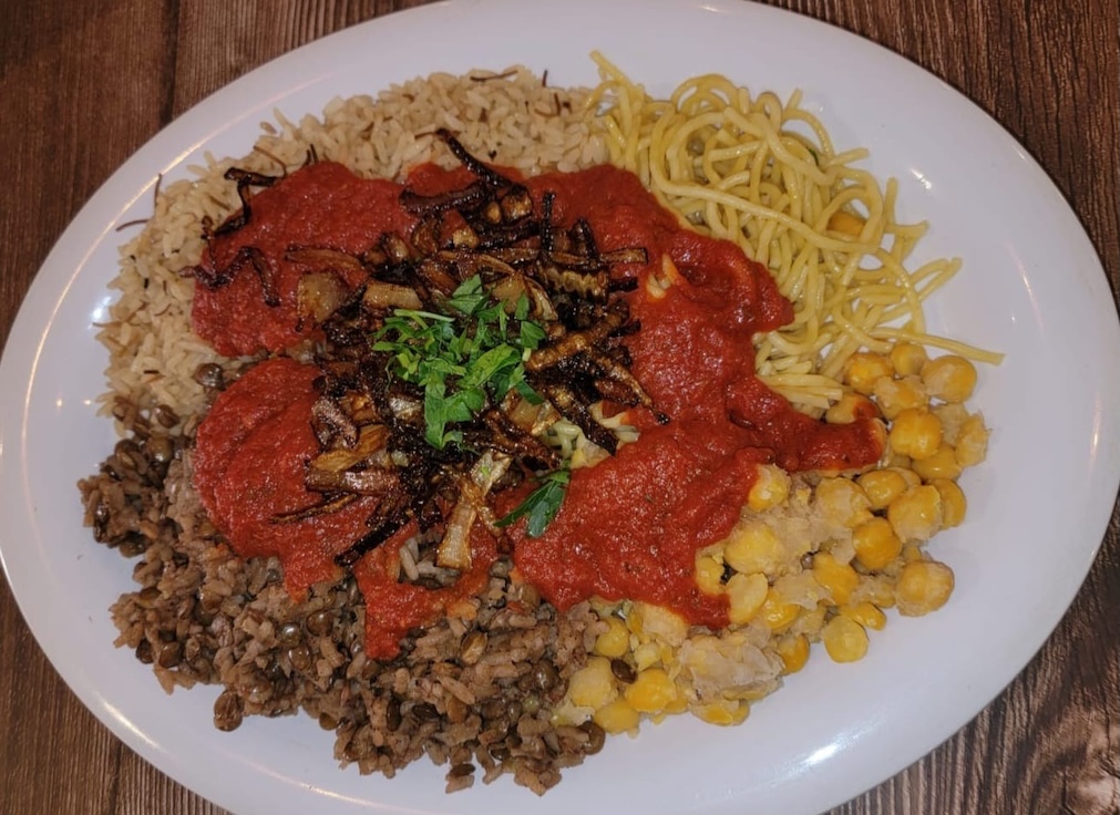 Plate with a serving of rice and lentils, yellow chickpeas, yellow noodles, topped with red tomato sauce and garnished with chopped parsley and fried onions on a wooden table.