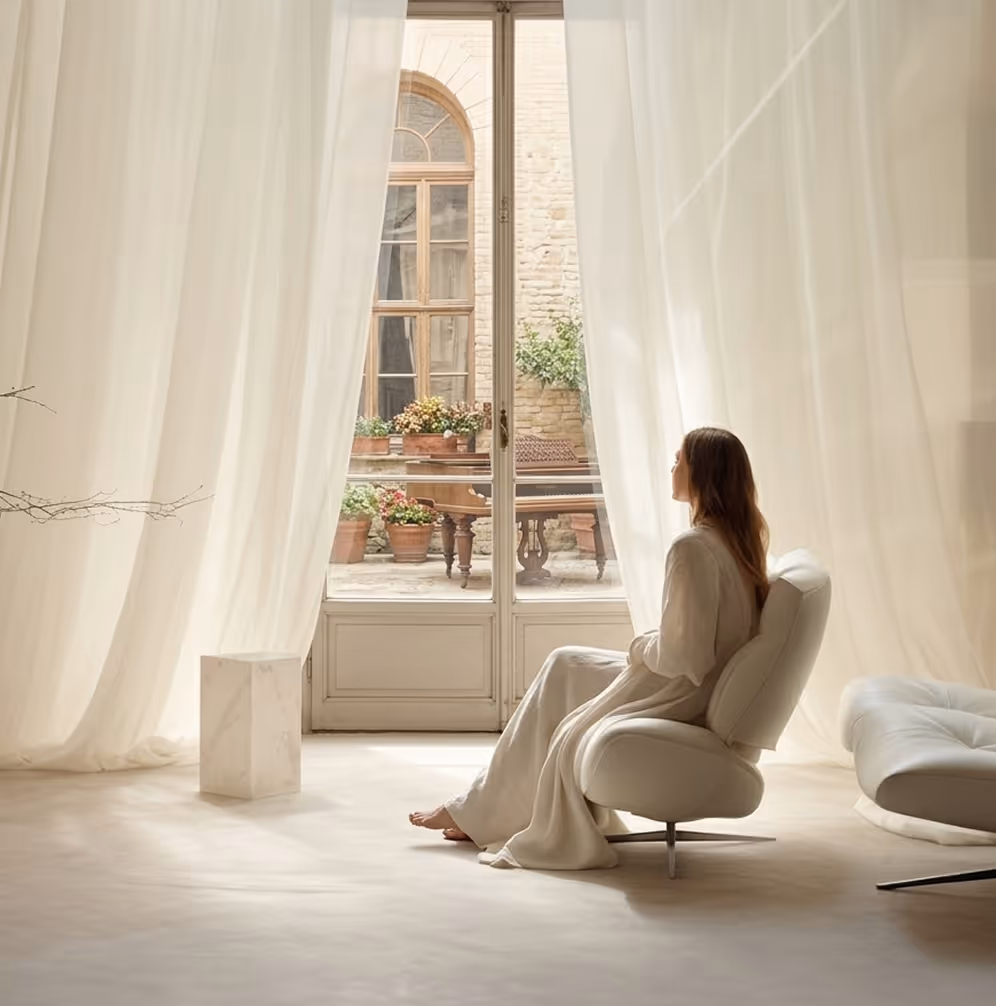 Woman in white dress sitting barefoot on a modern white chair by large glass doors with sheer white curtains and flower pots outside.