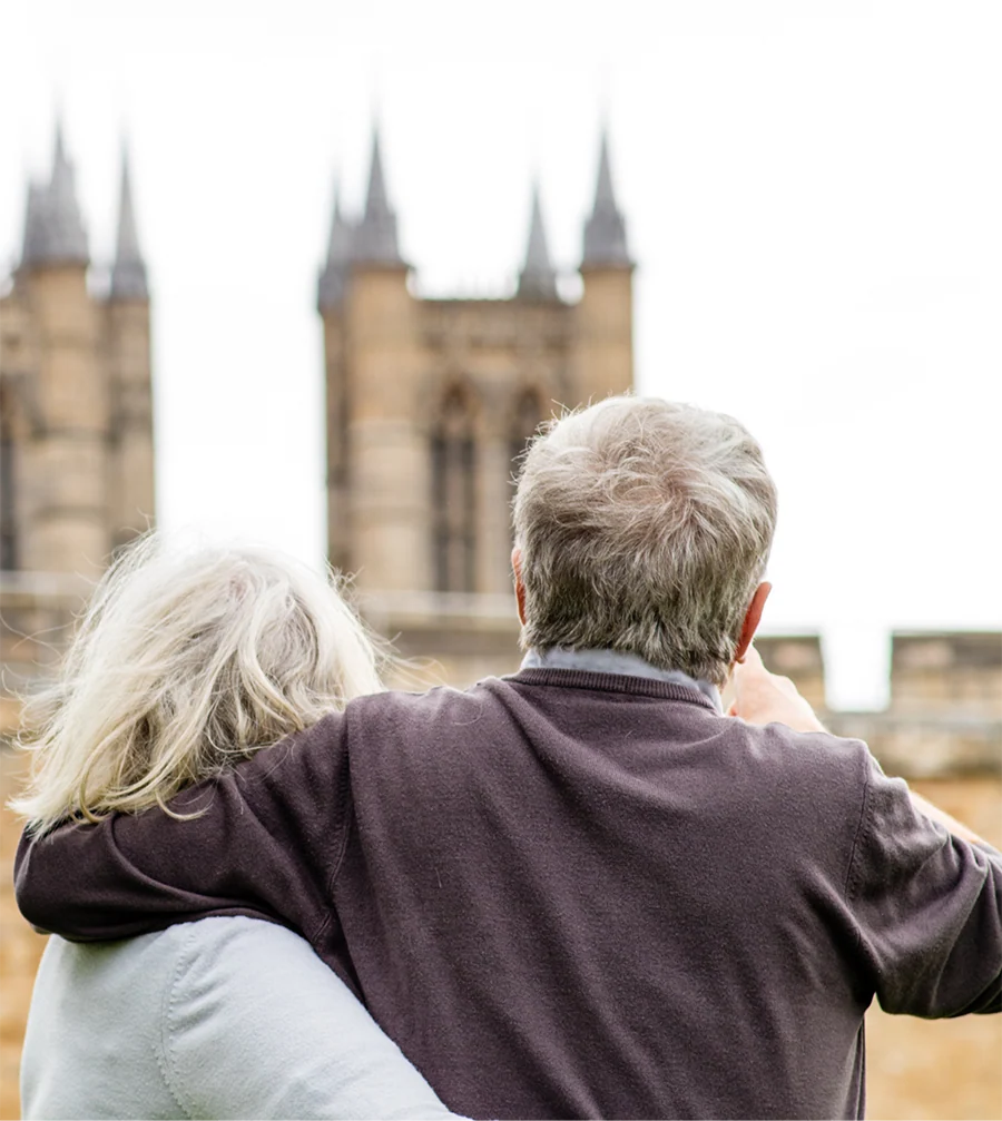 Older couple with gray hair embracing and looking at the towers of a historic stone building.