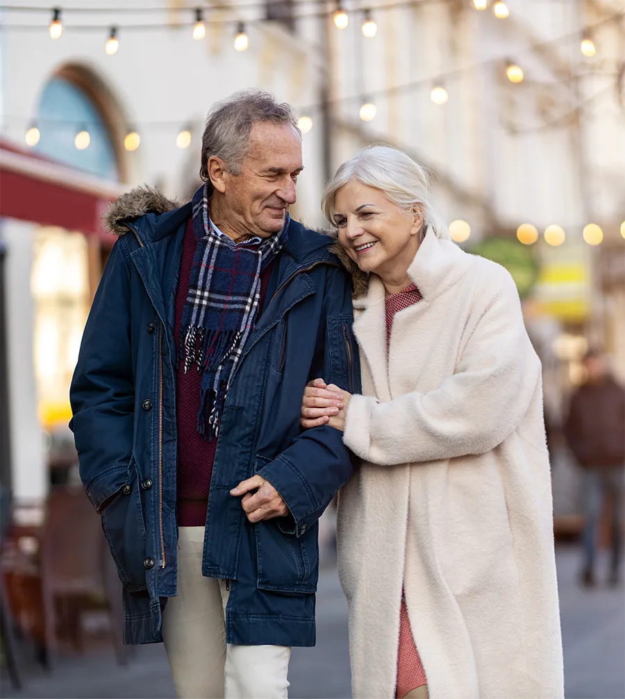 Smiling elderly couple walking arm in arm down a city street adorned with string lights.