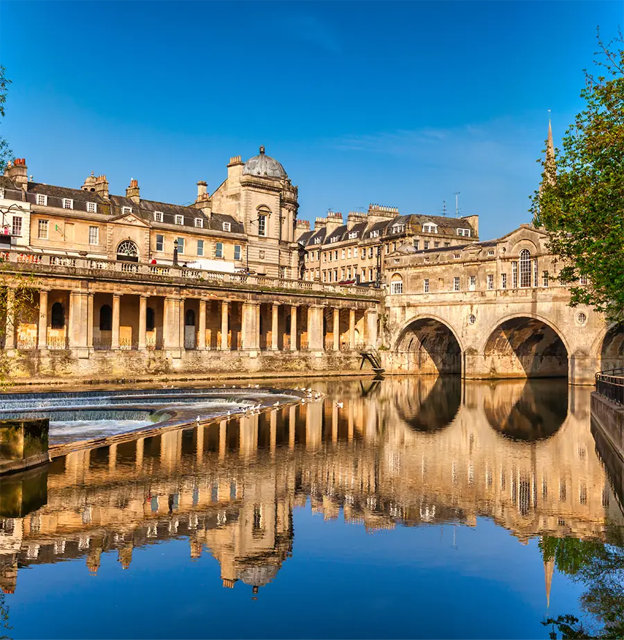 Historic stone buildings with columns and arches reflected in calm river water under clear blue sky.