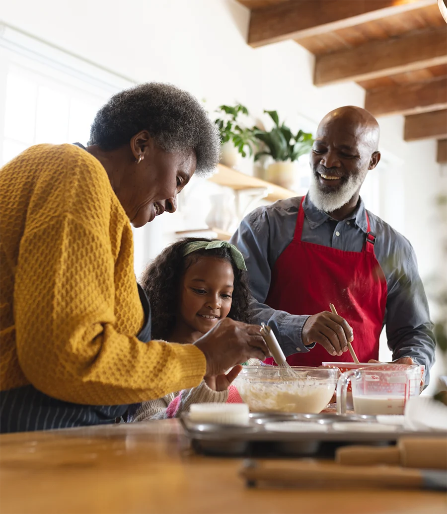 Elderly couple and a young girl happily mixing batter in a kitchen with baking cups and measuring cups nearby.