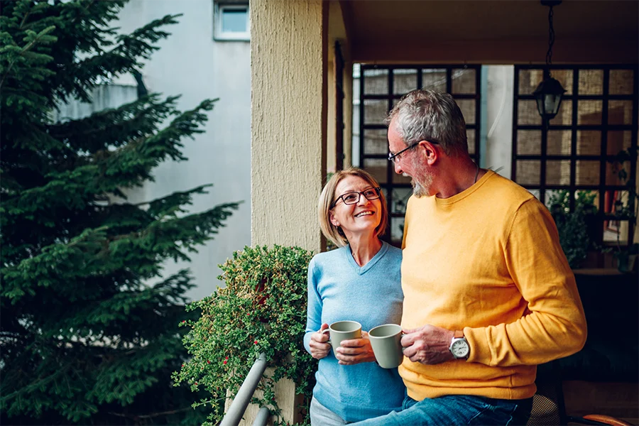 Smiling elderly couple holding coffee mugs, standing together on a balcony with greenery in the background.