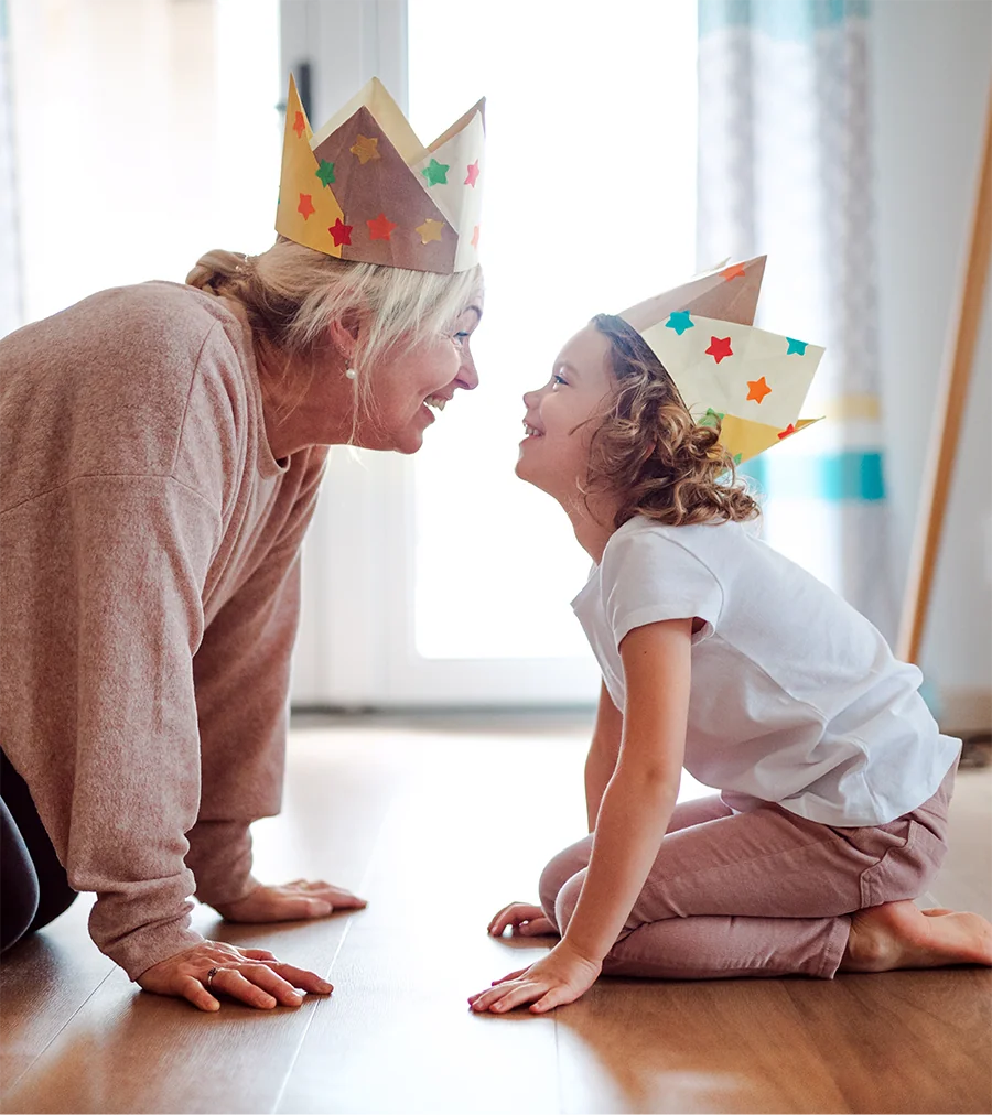 Older woman and young girl wearing paper crowns smiling and facing each other while kneeling on the floor.