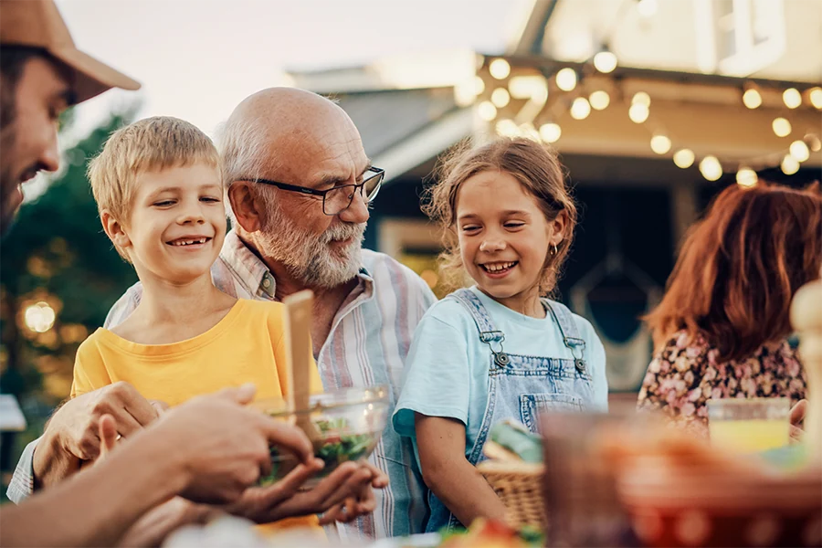 Smiling elderly man with glasses sitting outdoors between a young boy in a yellow shirt and a girl in denim overalls, sharing a joyful moment.