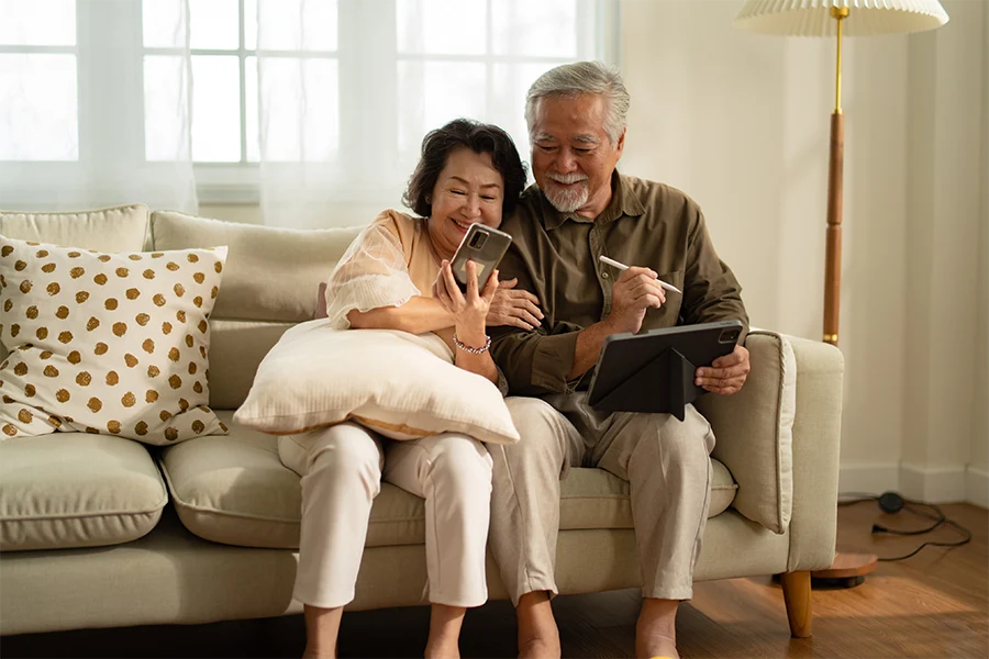 Senior couple sitting on a beige couch, smiling while looking at a smartphone and tablet in a cozy living room.