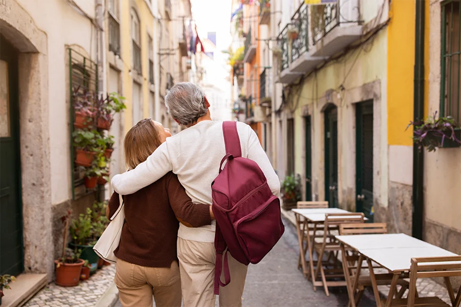 Older couple walking arm in arm down a narrow European street with colorful buildings and outdoor café tables.
